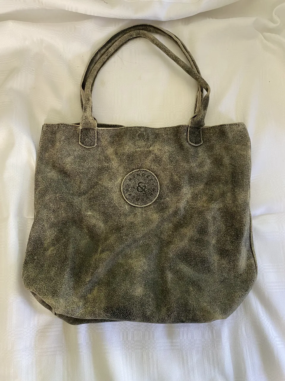 Bolso tote de cuero sintético con textura desgastada y detalle de logo circular metálico en el frente. Posee dos asas cortas.