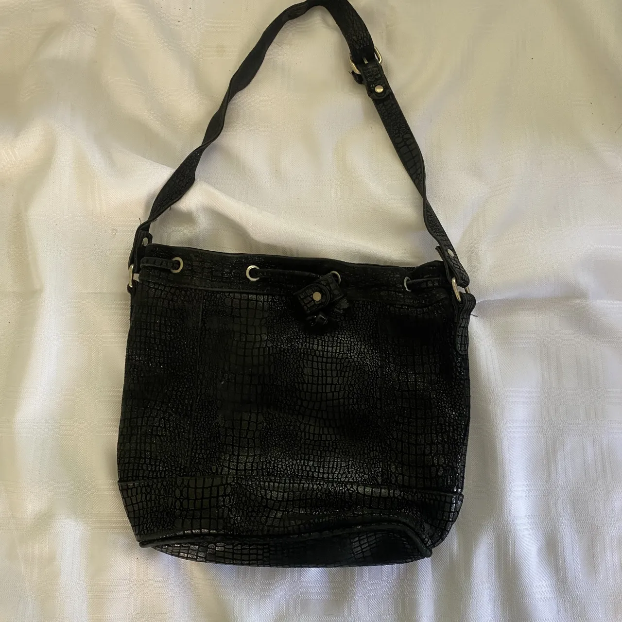 Bolso negro con textura de cocodrilo y cierre de cordón. Tiene una correa ajustable para llevar al hombro.