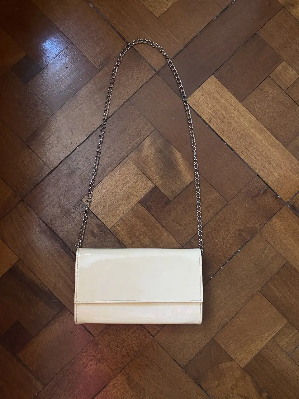 Cartera de mano estilo sobre, color crudo/nude, con cadena plateada. Símil charol. Cierra con botón imantado, tiene un pequeño bolsillo interno. Ideal para salidas o el día a día. 