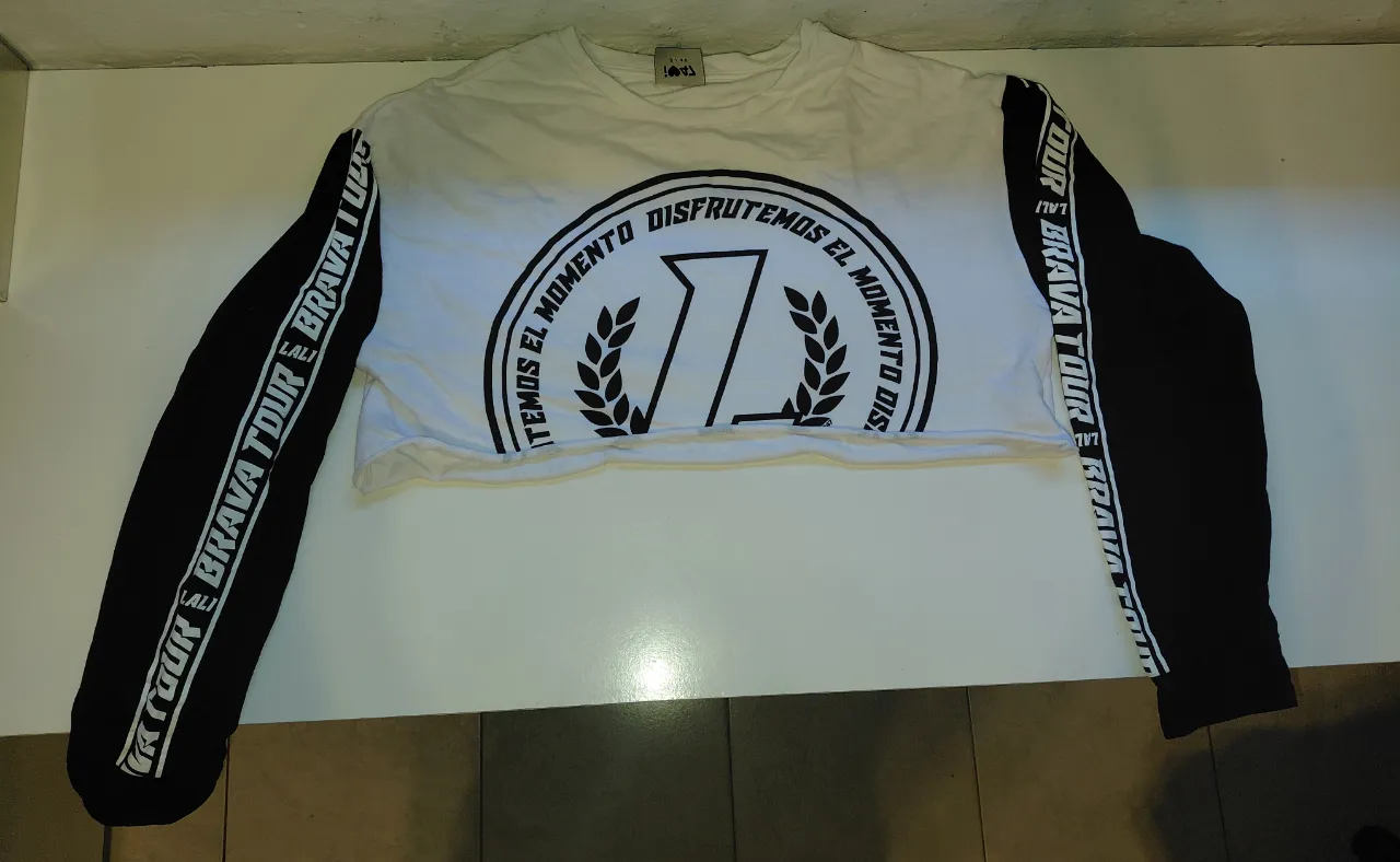 Remera crop top blanca con mangas largas negras. Tiene un estampado circular en el frente con la leyenda "EL MOMENTO DISFRUTEMOS EL MOMENTO" y un diseño central. Las mangas tienen una estampa lateral con la frase "BRAVA TOUR". Ideal para un look urbano y canchero.