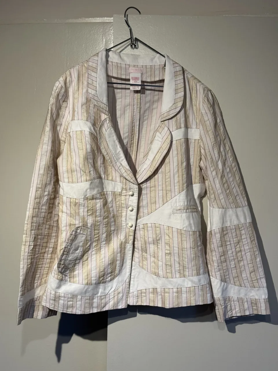 Blazer a rayas verticales en tonos blanco, rosa pálido y dorado. Posee cuello solapa, tres botones al frente y detalles de recortes en tela blanca. Mangas largas.