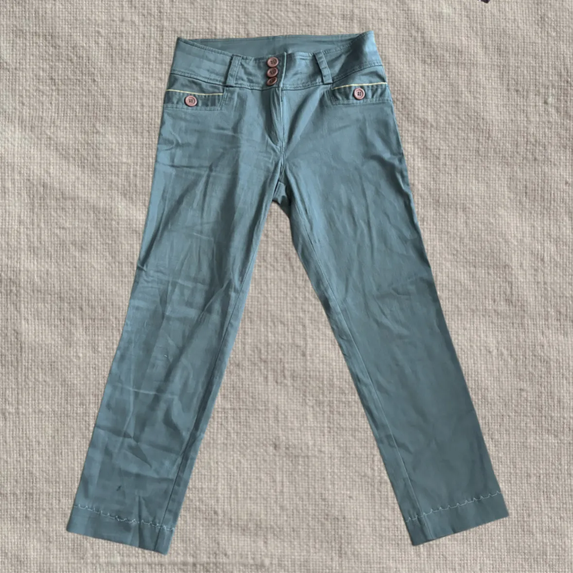 Pantalón de tela color gris con tres botones en la cintura y dos bolsillos delanteros con detalle de borde amarillo y botón. La botamanga tiene un pespunte decorativo.