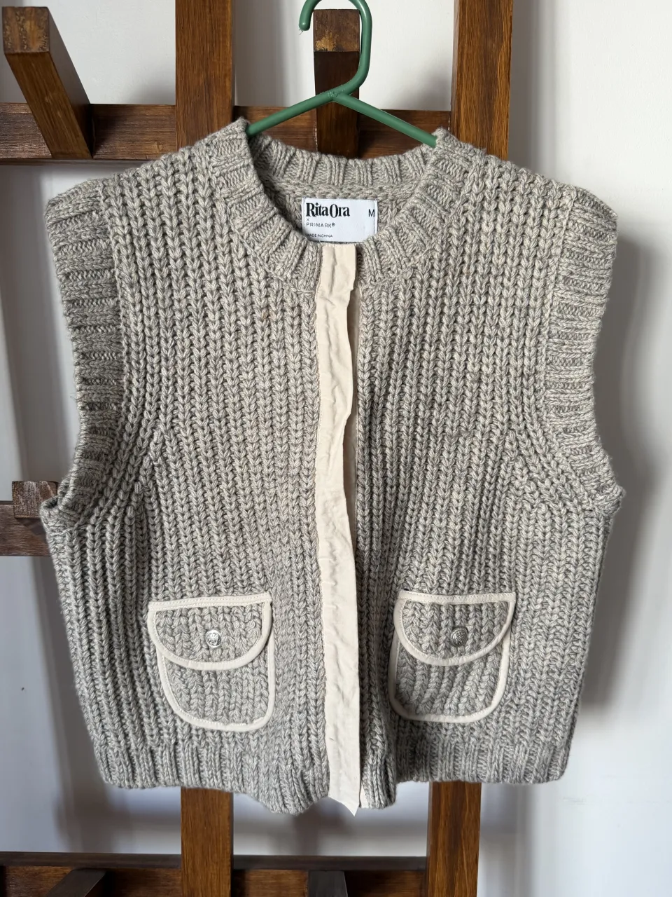 Chaleco tejido de punto grueso, color gris melange. Posee cuello redondo, dos bolsillos delanteros con solapa y botones plateados, y una tira central de tela color crudo con broches a presión. Ideal para un look casual y moderno.