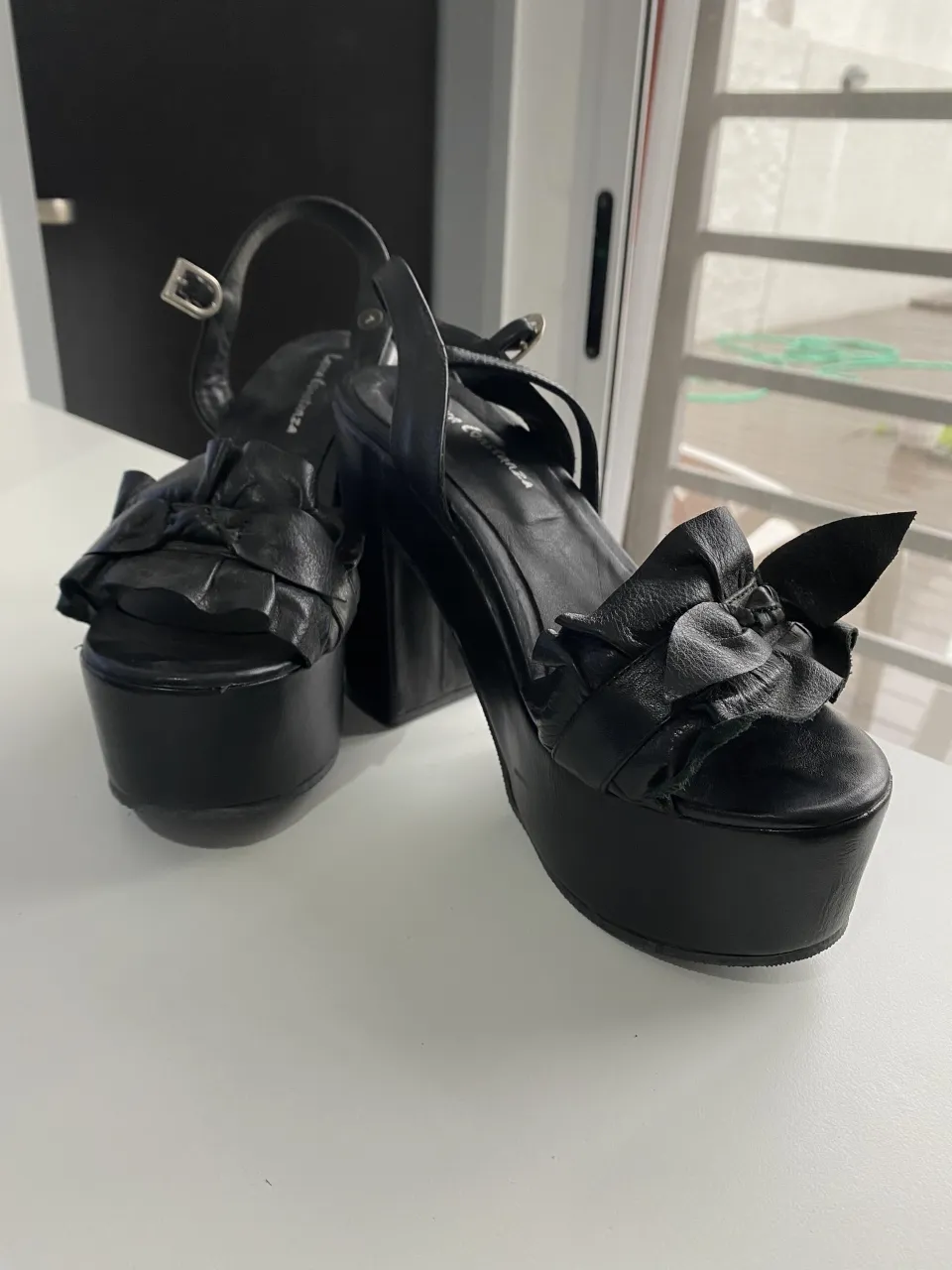 Sandalias negras con plataforma y taco alto. Tienen tiras finas en el empeine con detalle de volados y una tira que sujeta el tobillo con hebilla.