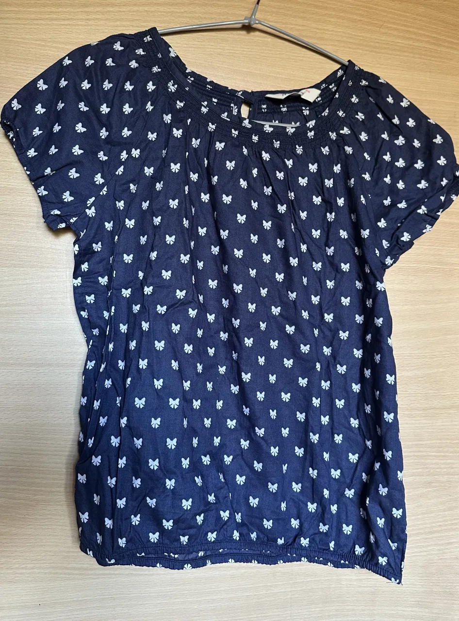 Blusa azul con estampa de moños blancos. Tiene cuello redondo con fruncido y mangas cortas. Ideal para un look casual y fresco.