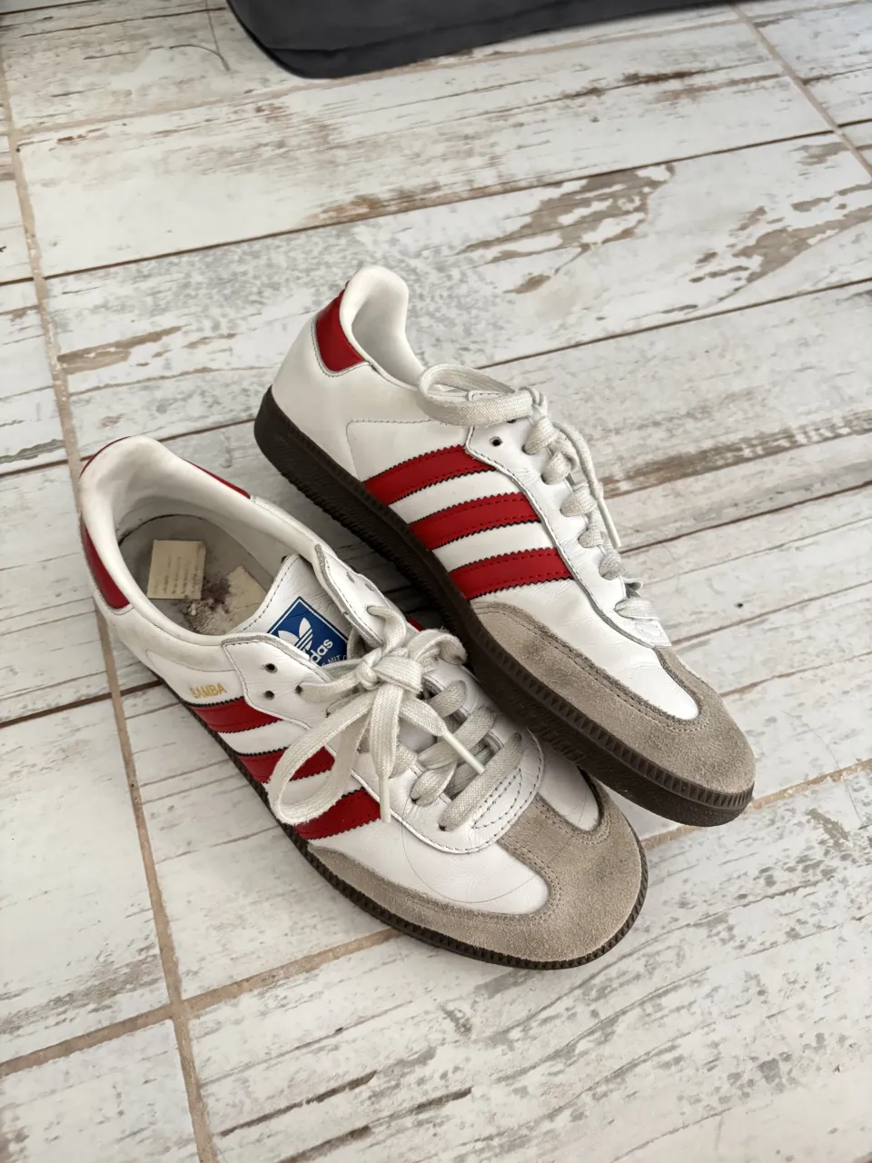 Zapatillas Adidas Samba blancas con detalles en rojo y gamuza gris. Cordones blancos y suela de goma oscura.