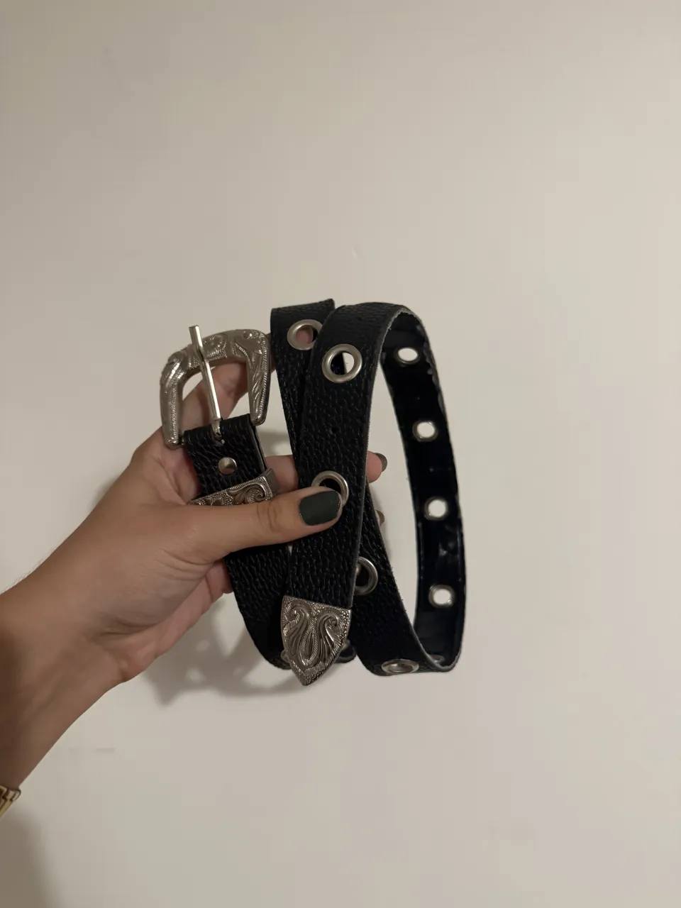 Cinturón negro con hebilla plateada y detalles grabados. Posee múltiples ojales metálicos a lo largo de la tira. LARGO APROX: 1m