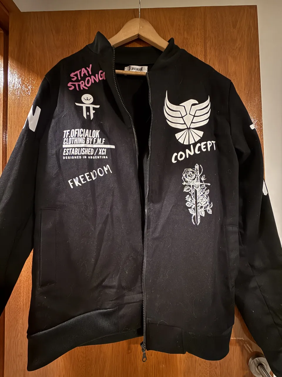 Campera bomber negra con inscripciones y estampas en blanco y rosa. En el frente tiene un águila con la palabra "CONCEPT" debajo y una rosa con una daga. En la manga izquierda tiene "STAY STRONG" y "FREEDOM". En la espalda tiene "LIVE FAST", "STAY STRONG", "DIE YOUNG" y "THINK MAKE MISTAKES".