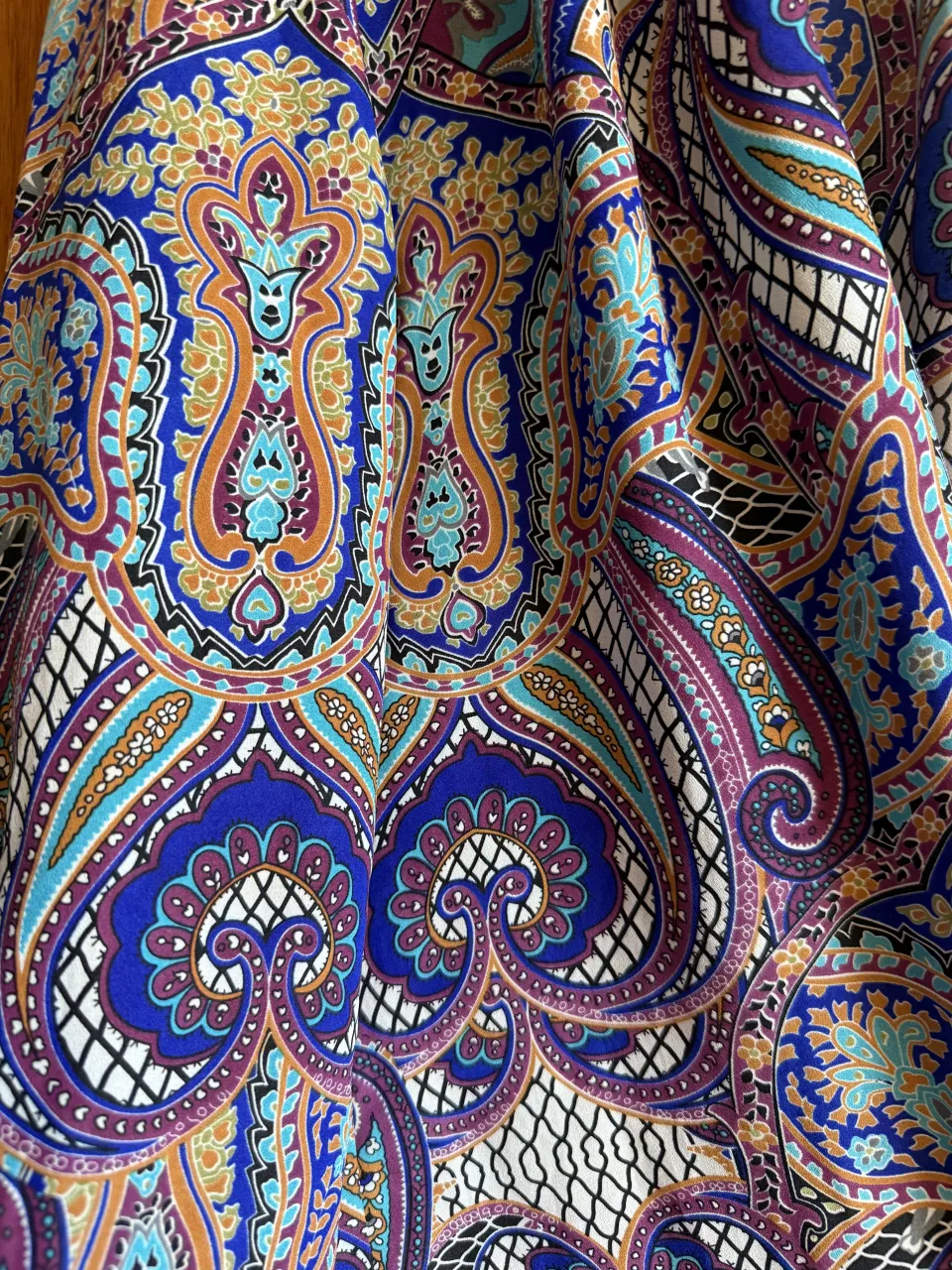 Blusa estampada con motivos paisley en tonos azules, violetas, naranjas y blancos. Posee cuello redondo con pequeña abertura y botones al frente. Las mangas son largas y amplias.