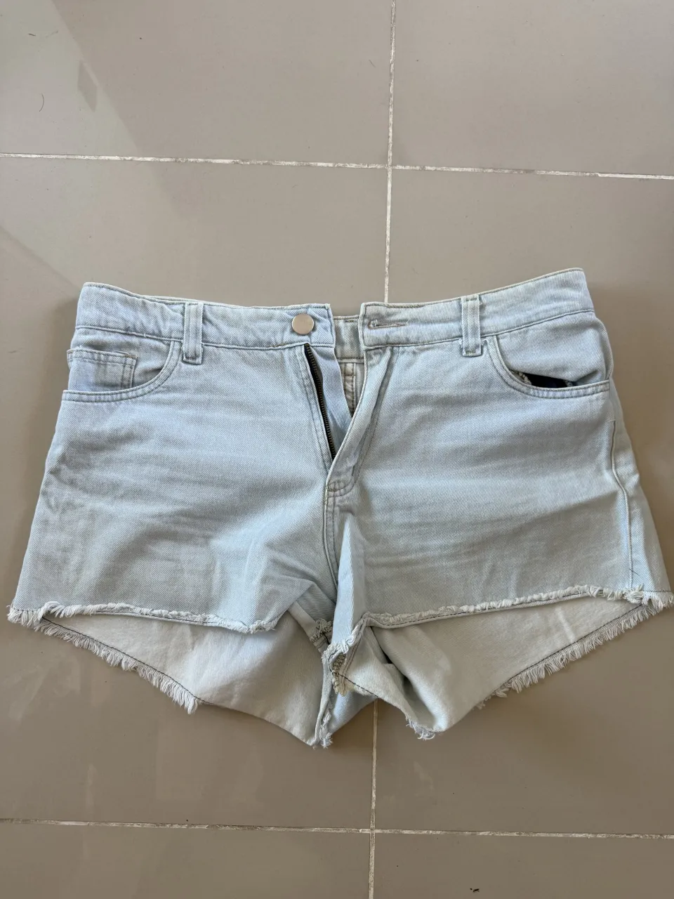 Shorts de jean tiro alto, color celeste claro, con roturas y flecos en el borde inferior. Cierre con botón y cierre.