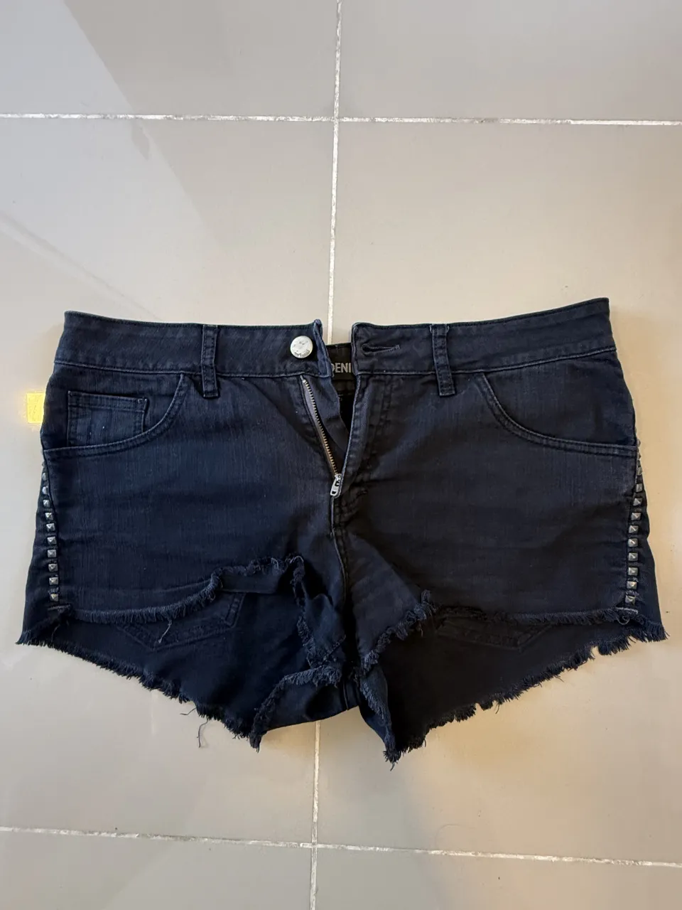 Shorts de jean negros con tachas plateadas en los laterales y ruedo deshilachado. Cierre y botón.