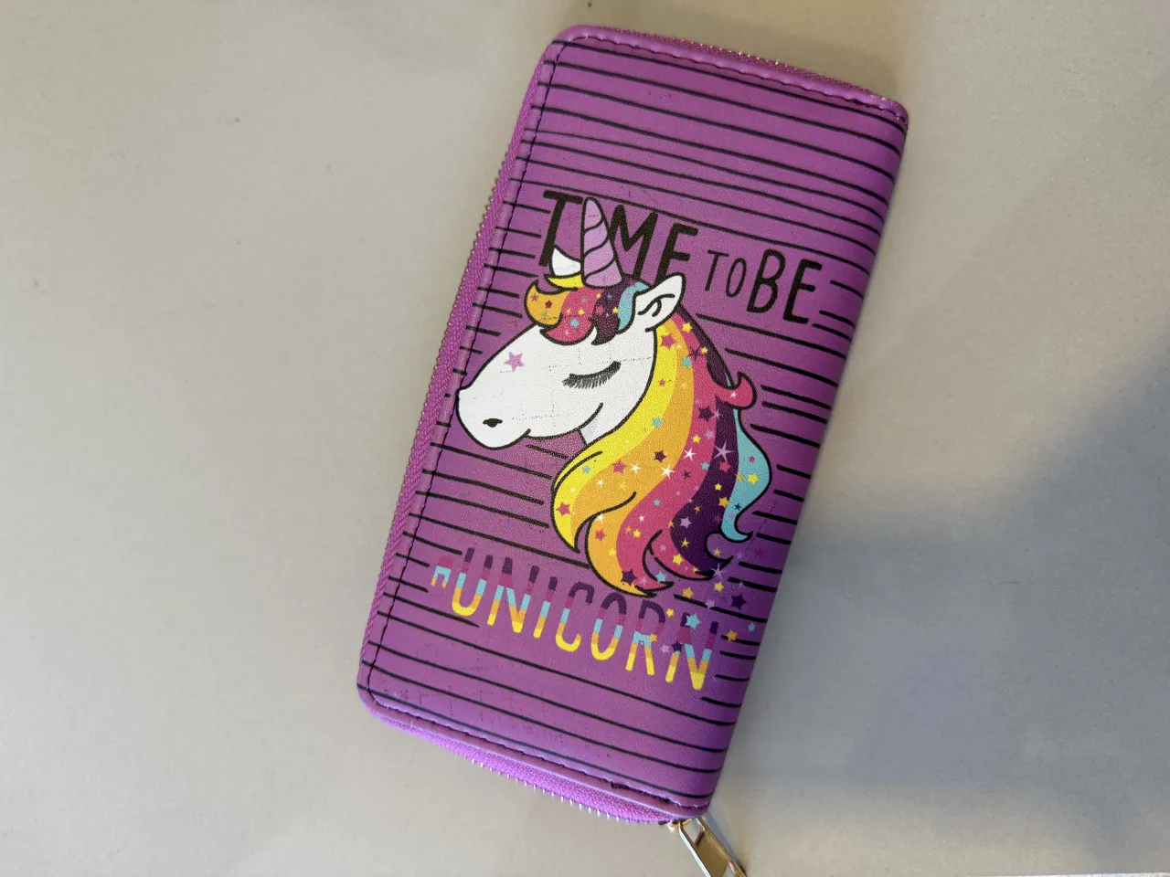 Billetera violeta con estampado de unicornio y la frase "TIME TO BE UNICORN". Tiene cierre y múltiples compartimentos internos.