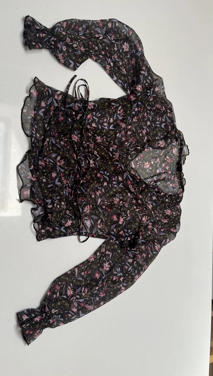 Blusa vaporosa con estampado floral en tonos oscuros y detalles de volados. Posee mangas largas acampanadas y un lazo para atar en la cintura, creando un efecto cruzado muy femenino. Ideal para un look romántico y chic.