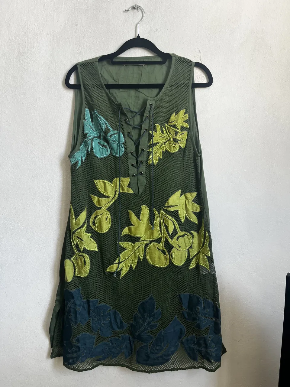 Vestido verde con detalles de hojas y flores bordadas en tonos verde lima y turquesa. Posee un diseño de red en la parte superior y cordones para atar en el escote. Ideal para un look fresco y bohemio.