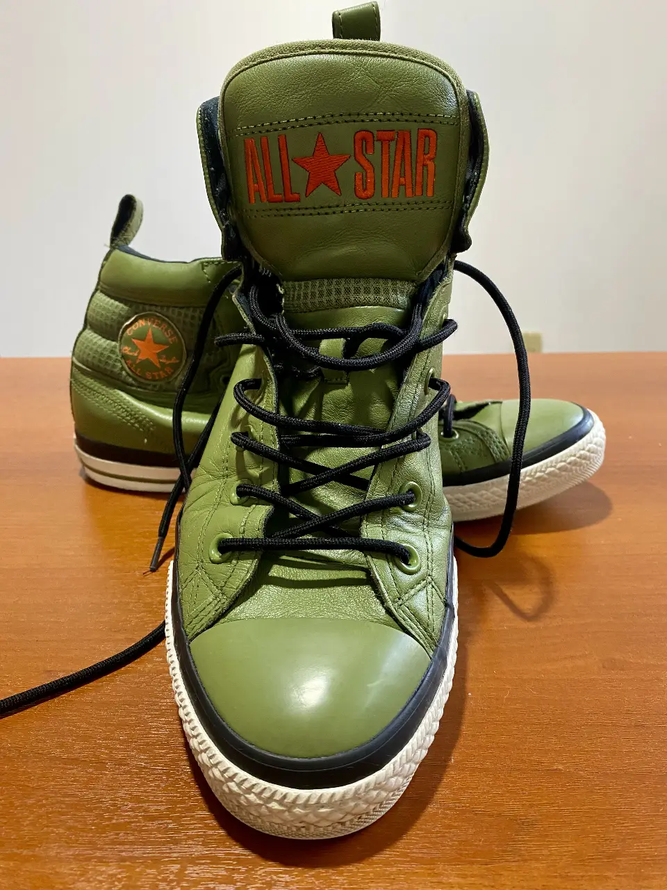 Zapatillas Converse All Star High Top Foot Locker Exclusive, de caña alta en color verde militar. Con detalles en naranja y negro, estas zapatillas son ideales para un look urbano y moderno. Tienen cordones negros y suela de goma blanca.