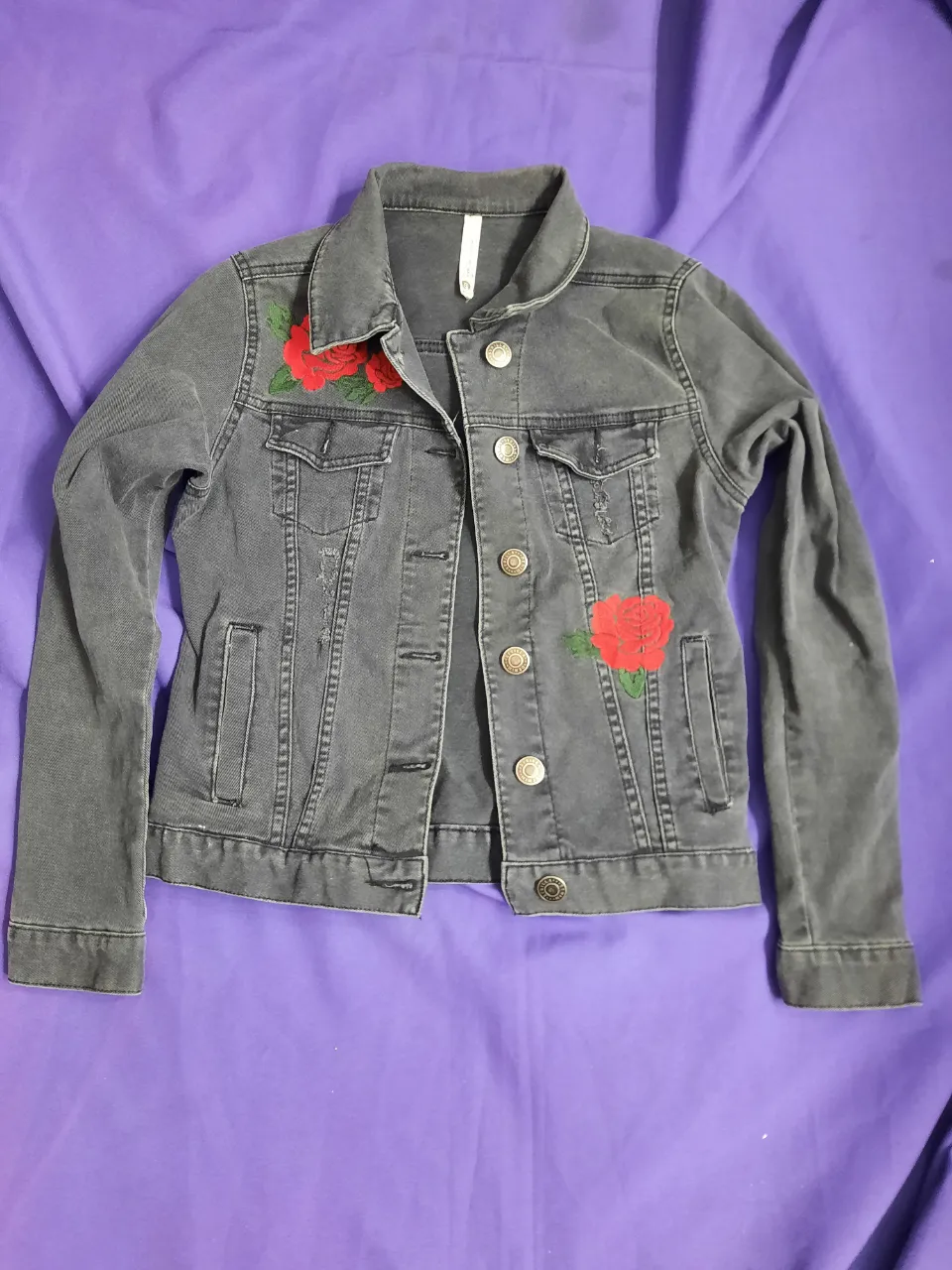 Campera de jean gris con detalles de flores rojas bordadas en la parte delantera. Posee botones metálicos y bolsillos por dentro y fuera.