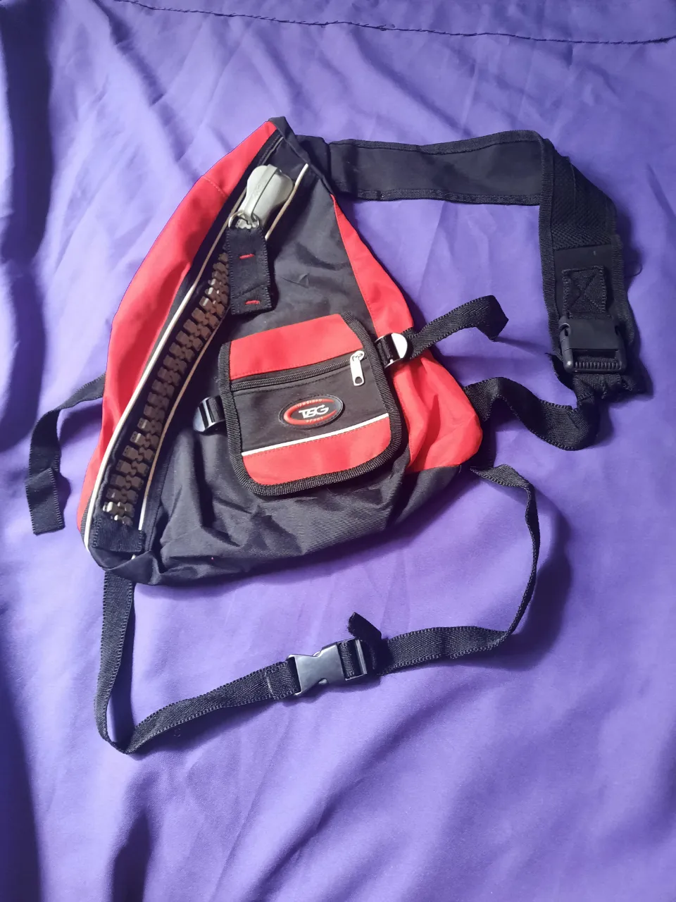Mochila urbana de tela negra y roja con cierre principal grande y bolsillo frontal con cierre. Correa ajustable. Con forma de guitarra pequeña.