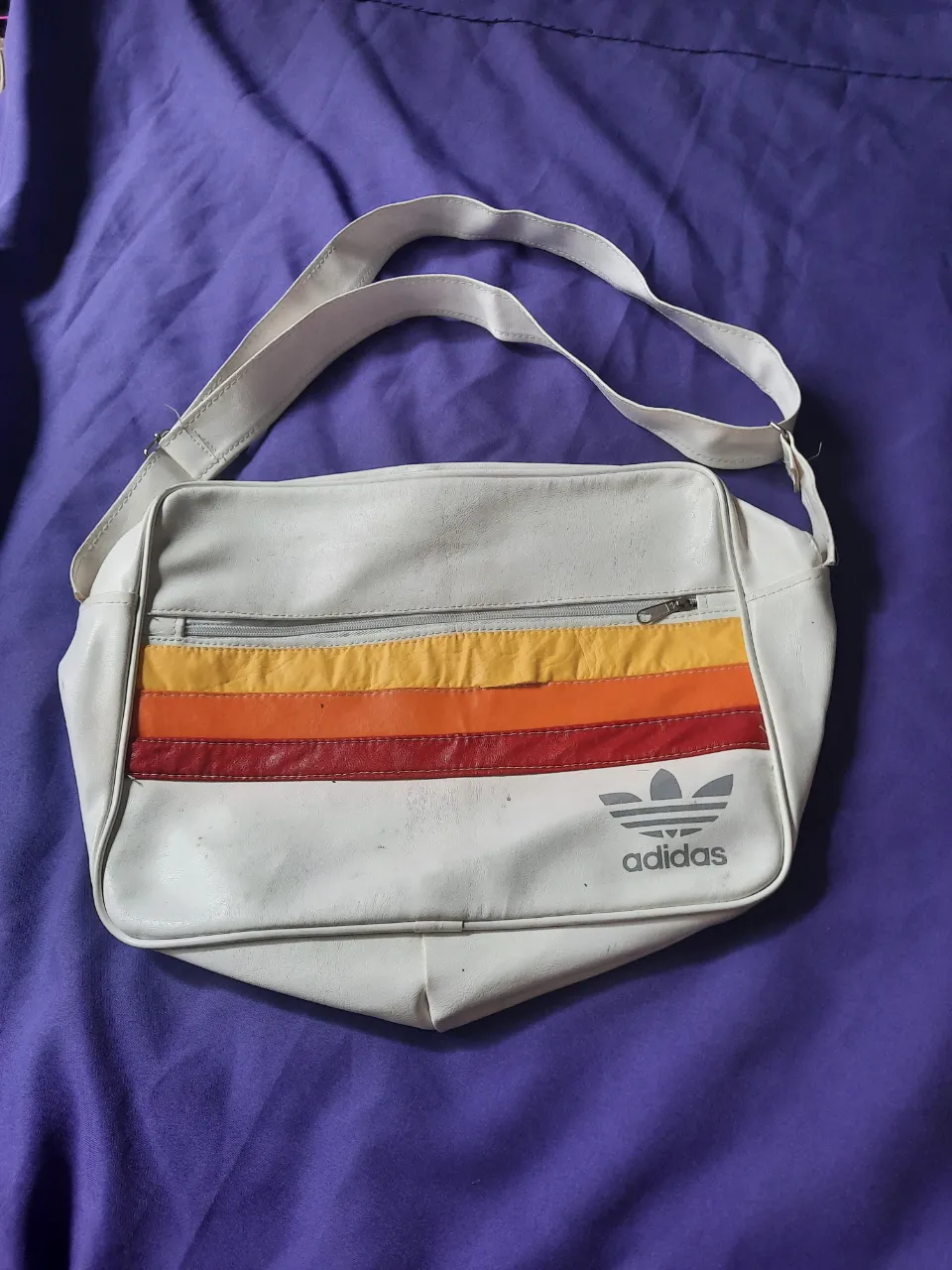 Bolso blanco de cuero sintético con tres franjas horizontales de colores (amarillo, naranja y rojo) en la parte frontal. Posee un bolsillo con cierre en la parte superior y el logo de Adidas estampado en gris. El interior está forrado en tela. Tiene algunos desperfectos que se pueden ver en las fotos, pero no afecta en el uso.