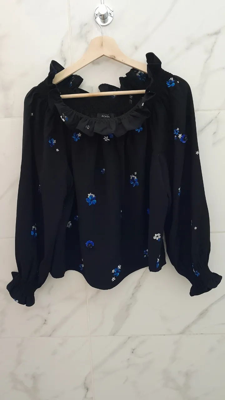 Blusa negra con mangas largas y volados en el cuello y puños. Posee un estampado floral en tonos azules y blancos. El cuello es escotado y fruncido.