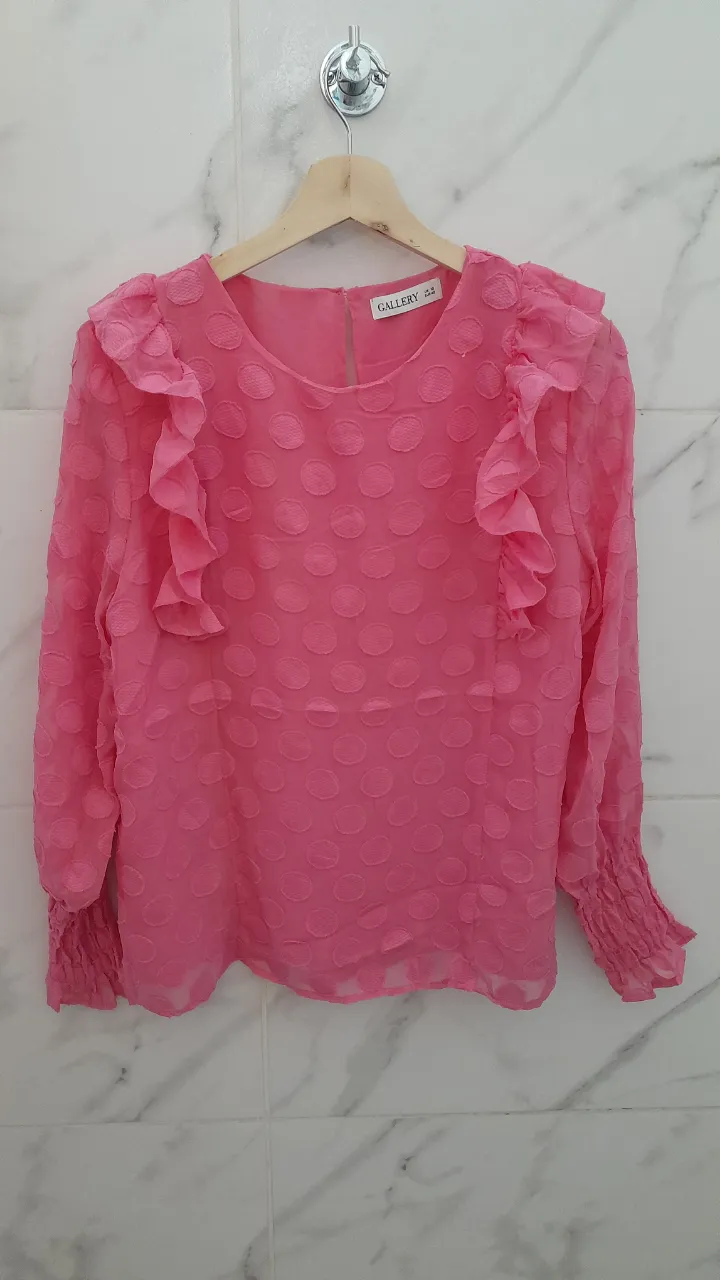 Blusa rosa con mangas largas y volados en los hombros. Posee puños elastizados con detalle de fruncido. La tela tiene un relieve de lunares.