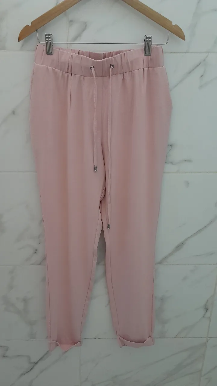 Pantalón rosa claro con cintura elastizada y cordón ajustable. Posee bolsillos laterales y traseros. Los bajos están doblados.