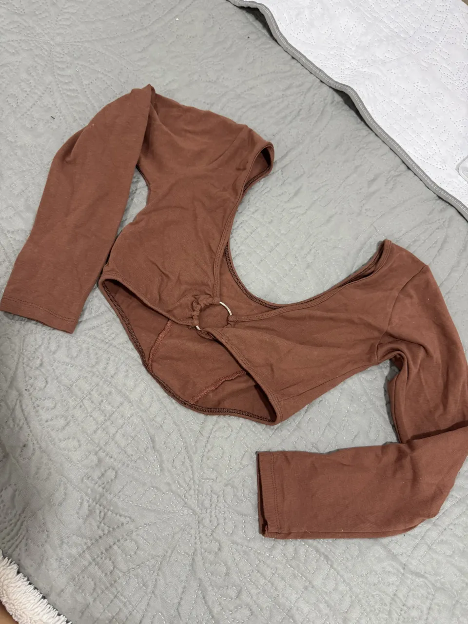 Remera manga larga marrón con escote pronunciado y detalle de aro metálico en el centro. Posee costuras marcadas en el frente y puños en las mangas.