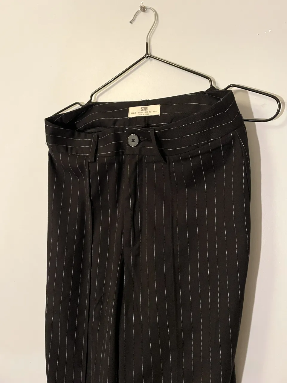Pantalón de vestir negro con finas rayas blancas verticales. Diseño clásico y elegante, ideal para un look formal o semi-formal. Posee un botón en la cintura y cierre. Tiene un dobladillo que se puede descoser.