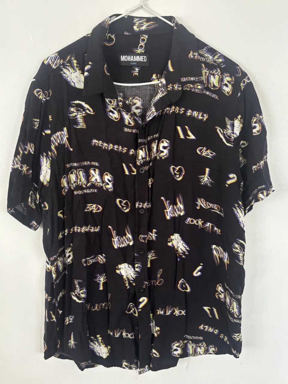 Camisa negra de manga corta con estampado integral de letras y figuras abstractas en colores blanco, amarillo y violeta. Cuello camisero y botones frontales.