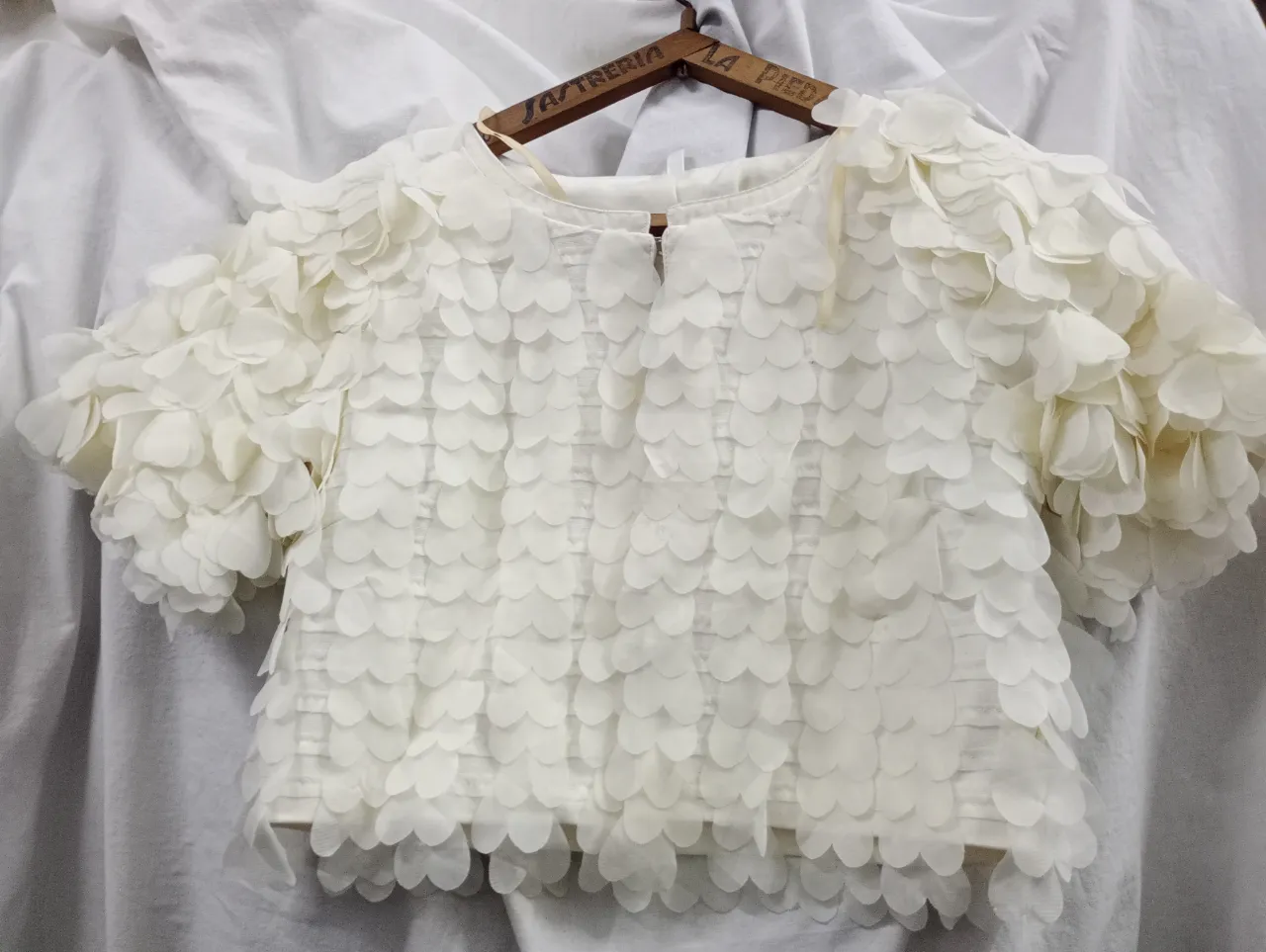 Blusa blanca con mangas cortas y apliques de pétalos en toda la prenda. Posee un corte a la cintura. se abrocha con un solo broche, es una casaca corta. el color es blanco manteca 