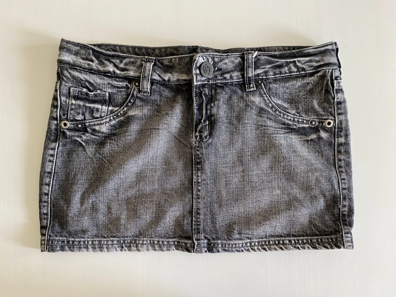 Pollera de jean gris desgastado. Tiene bolsillos delanteros y traseros. Los bolsillos traseros tienen cierre metálico plateado. En la parte trasera superior tiene un aplique bordado con alas, una corona y una cadena colgante.