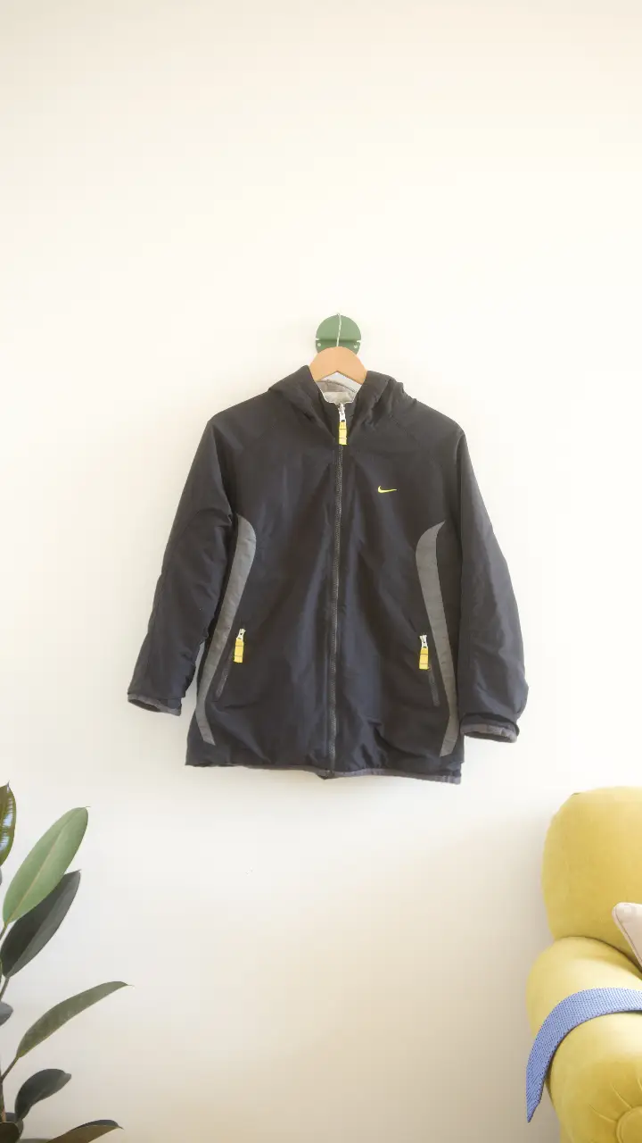 Campera deportiva Nike negra con detalles en gris y cierre. Logo amarillo en el pecho y nombre de la marca en la espalda. Ideal para actividades al aire libre o uso diario. Reversible. medidas largo 60 cm, ancho 45cm, mangas 50 cm