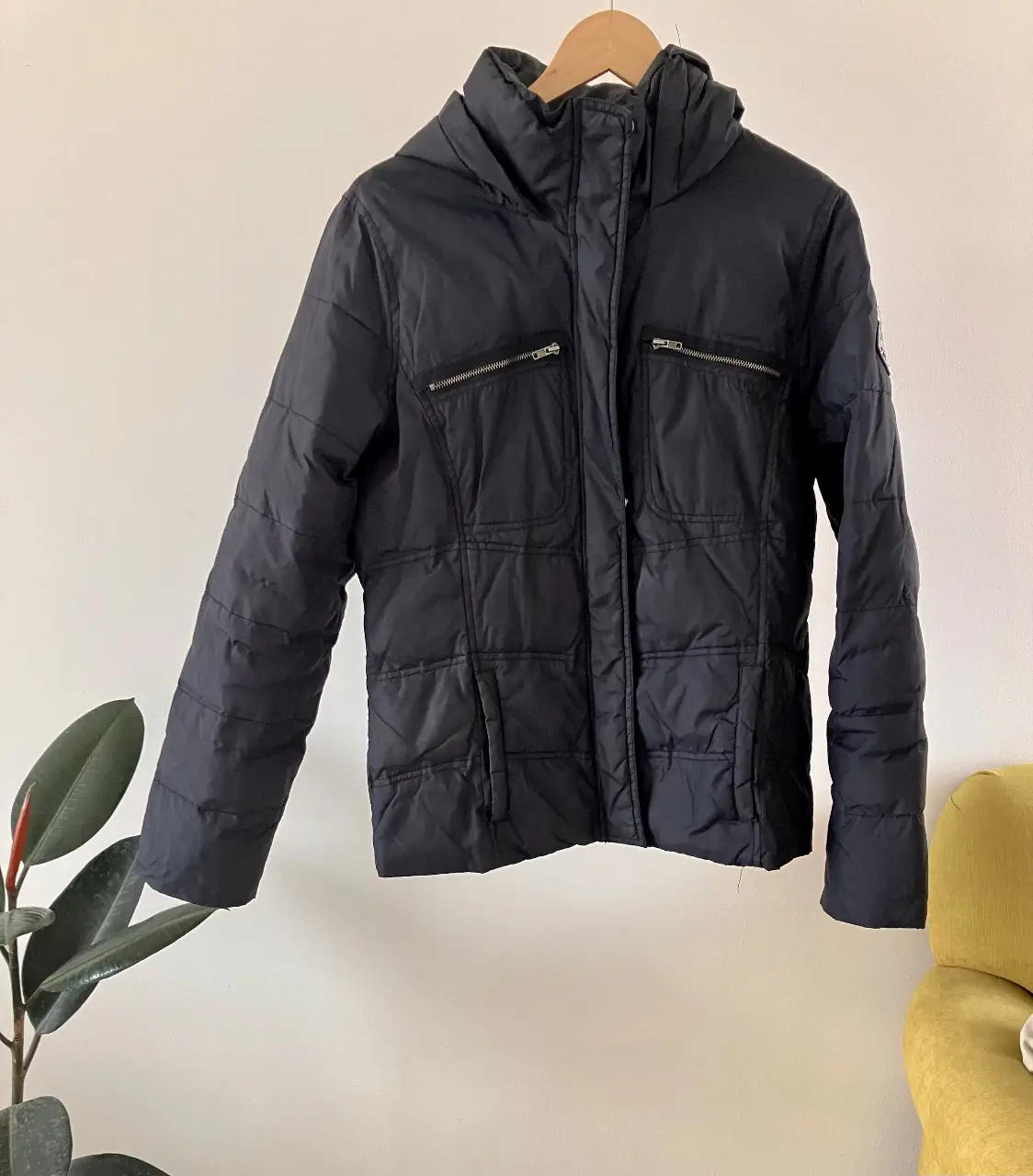 Campera negra acolchada con capucha, ideal para el invierno. Cuenta con cierre frontal, bolsillos con cierre y un parche decorativo en la manga. Perfecta para un look casual y abrigado.