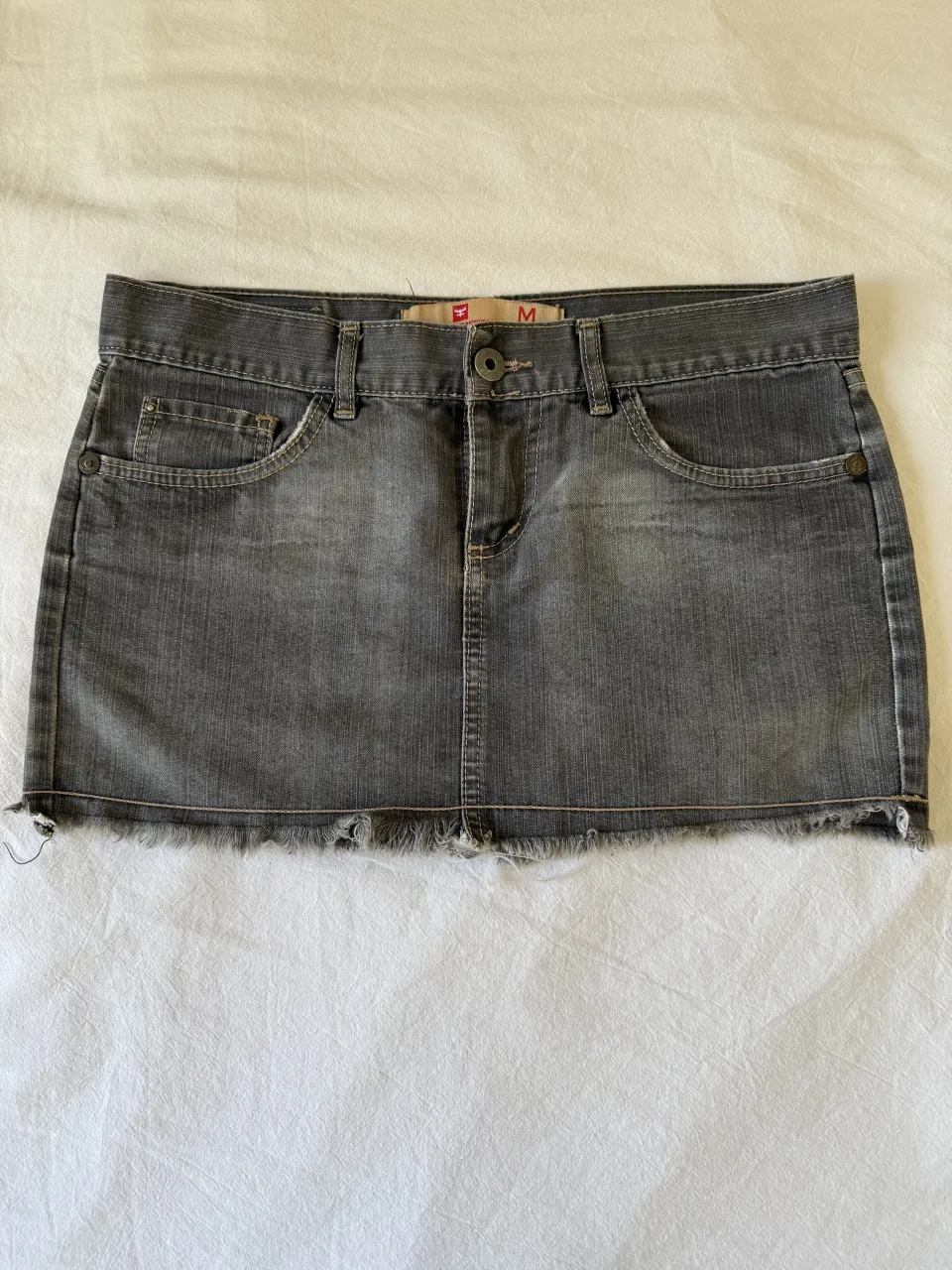 Pollera de jean gris con roturas en el borde inferior. Tiene bolsillos delanteros y traseros con cierre. Es más tirando a un S 