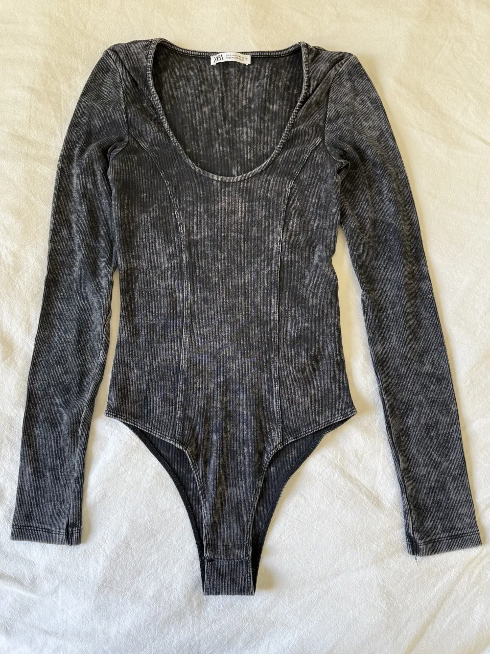 Body de manga larga con cuello escote en V y textura acanalada. Posee un estampado jaspeado en tonos de gris.