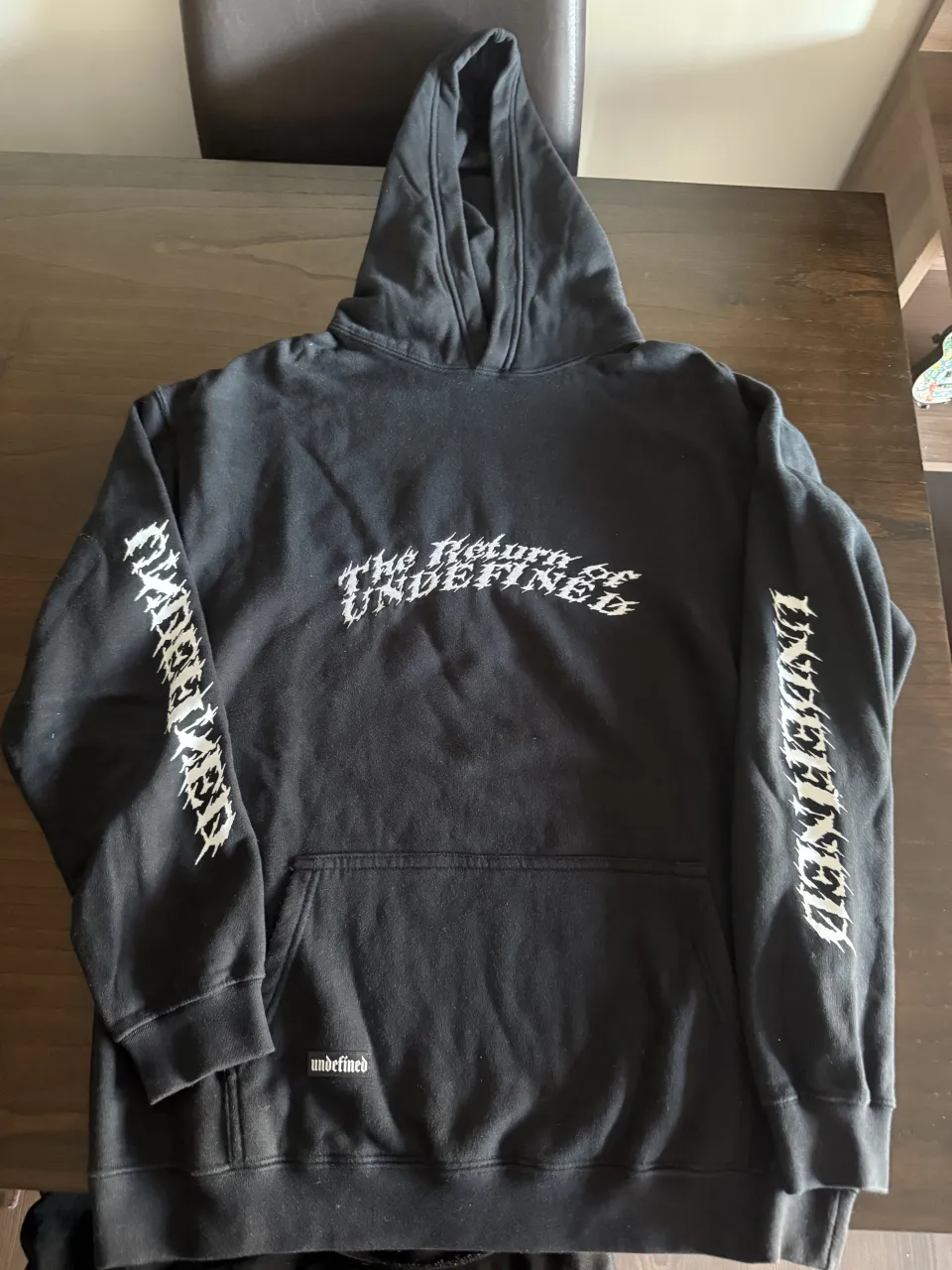 Buzo negro con capucha y estampa "The Return of UNDEFINED" en el frente y en las mangas. La estampa en la espalda es gráfica y abstracta con texto. Frizado.