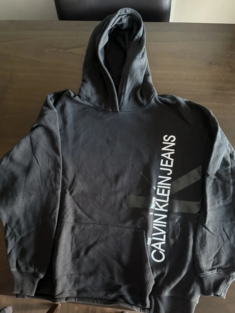 Buzo negro con capucha y bolsillo canguro. Estampado vertical con logo "CALVIN KLEIN JEANS" en blanco y logo de la marca en gris oscuro.