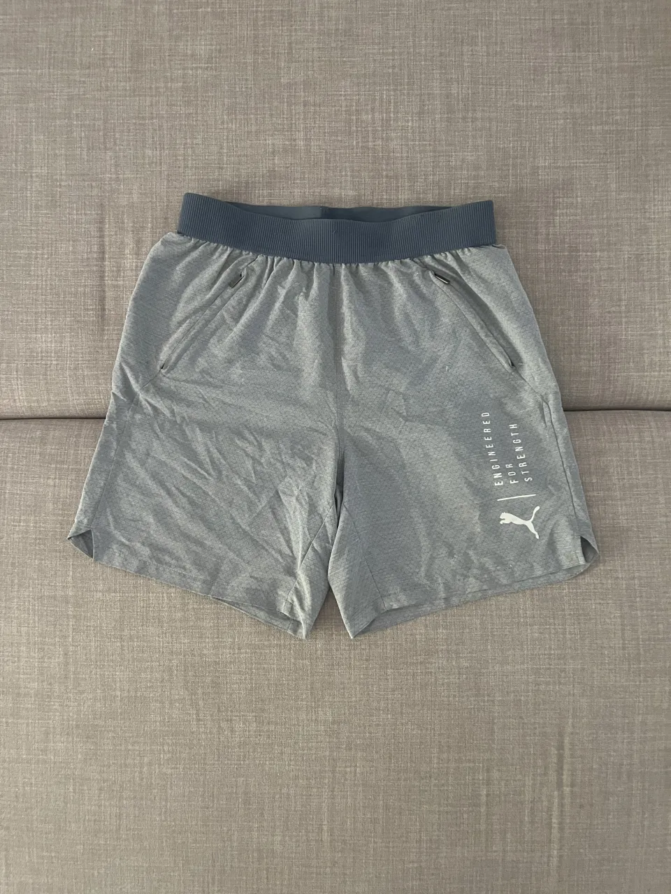 Shorts deportivos grises con logo de Puma y estampa "ENGINEERED FOR STRENGTH". Cintura elastizada con cordón interno y bolsillos laterales con cierre. Bolsillo trasero con cierre.