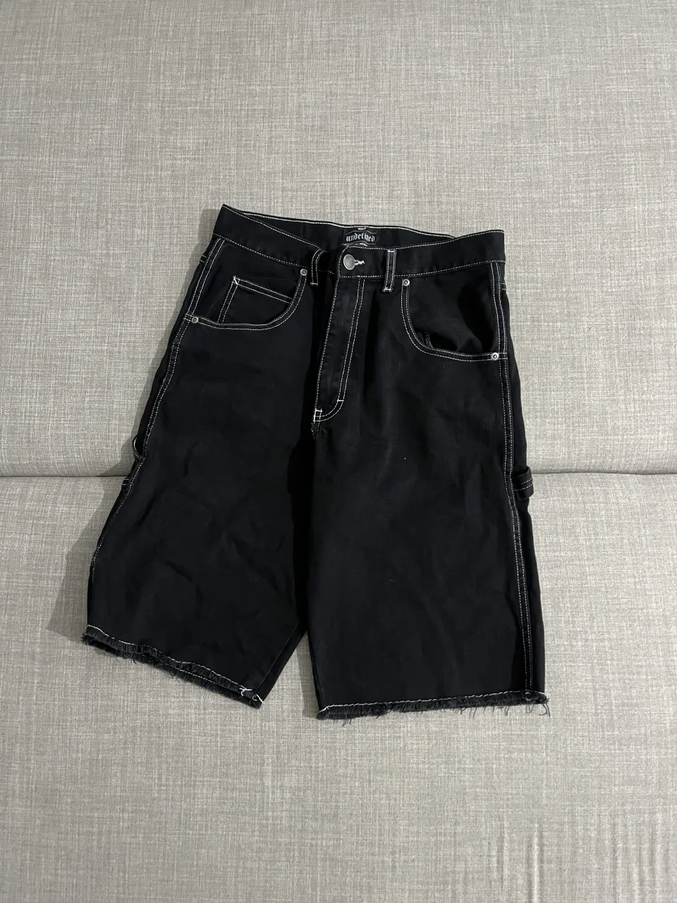 Shorts de jean negros con costuras blancas al tono. Poseen bolsillos delanteros y traseros, además de un bolsillo adicional tipo cargo en el lateral izquierdo. El borde inferior es deshilachado.