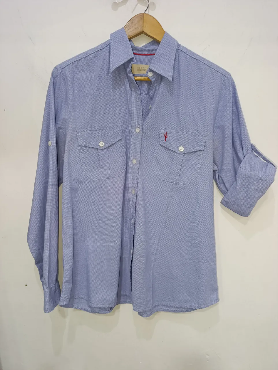 Camisa de manga larga de color celeste con pequeños detalles en blanco. Posee dos bolsillos en el frente, uno de ellos con un bordado rojo en forma de cactus. Los puños de las mangas son anchos y se pueden enrollar.