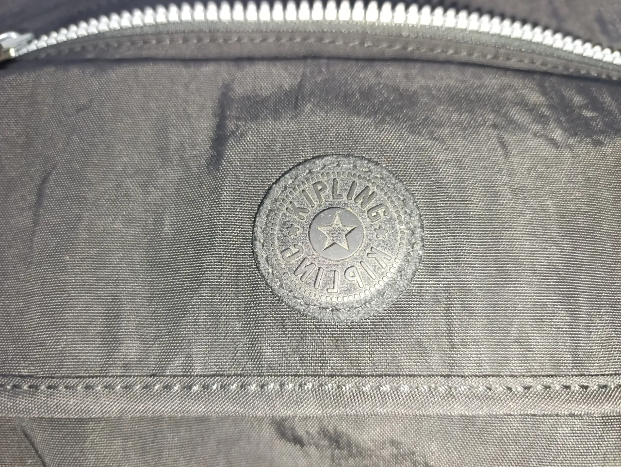 Cartera negra de tela con bolsillo frontal y cierre superior. Posee un logo circular de la marca en el frente y una etiqueta interna con el nombre de la marca y un dibujo de un mono.