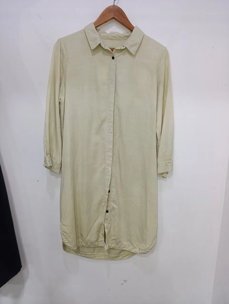 Camisa larga de jean color crudo con botones al frente y cuello de camisa. Las mangas son largas y se pueden ajustar con un botón.