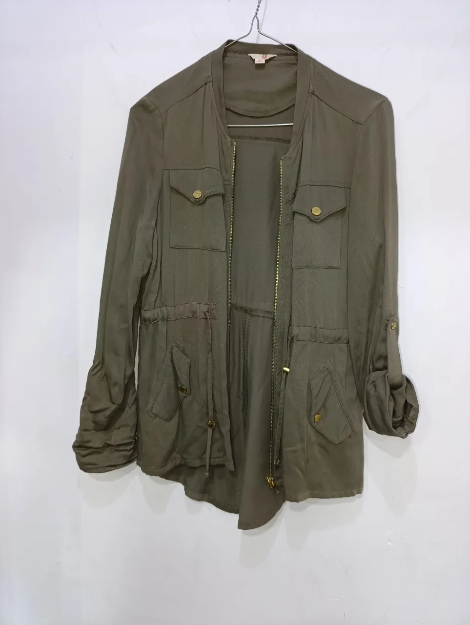 Chaqueta de color verde militar marca GUESS , con cuello redondo, cierre al frente y bolsillos con solapa y botones dorados. Posee cordón ajustable en la cintura y puños con detalle de botones.