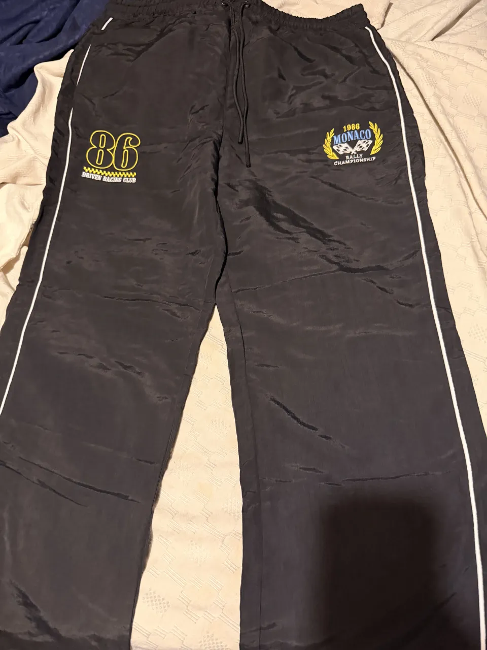 Pantalón de acetato negro con detalles de rayas blancas y amarillas en los laterales. Presenta bordados de "86 DRIVEN RACING CLUB" en amarillo y "1986 MONACO RALLY CHAMPIONSHIP" con laureles y banderas a cuadros en azul y amarillo.