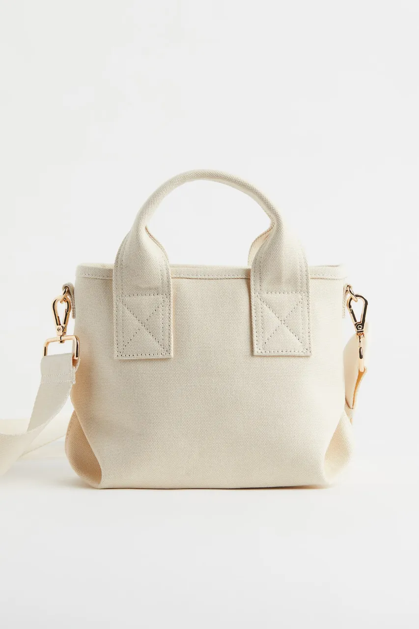 Bolso tote de tela color crudo con manijas cortas y correa larga desmontable. Tiene herrajes dorados.