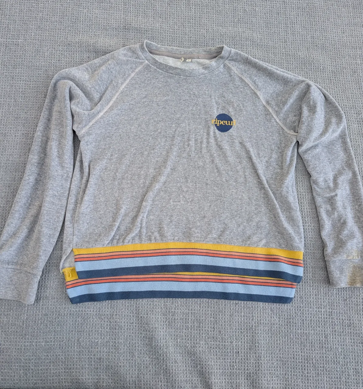 Buzo gris con cuello redondo y mangas largas. Presenta un detalle de franjas horizontales en la parte inferior del frente en tonos amarillo, naranja, celeste y azul oscuro. Posee un pequeño logo circular en el pecho izquierdo.