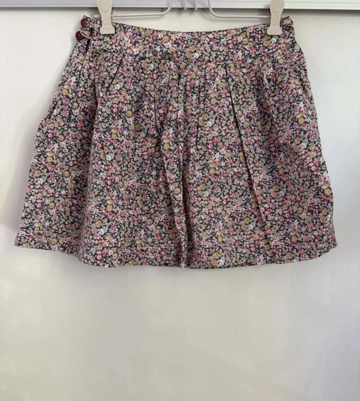 Pollera corta estampada con flores pequeñas en tonos rosas, amarillos y blancos sobre fondo oscuro de 47 street. Tiene tablas  y cierre lateral con botones decorativos en la parte superior.