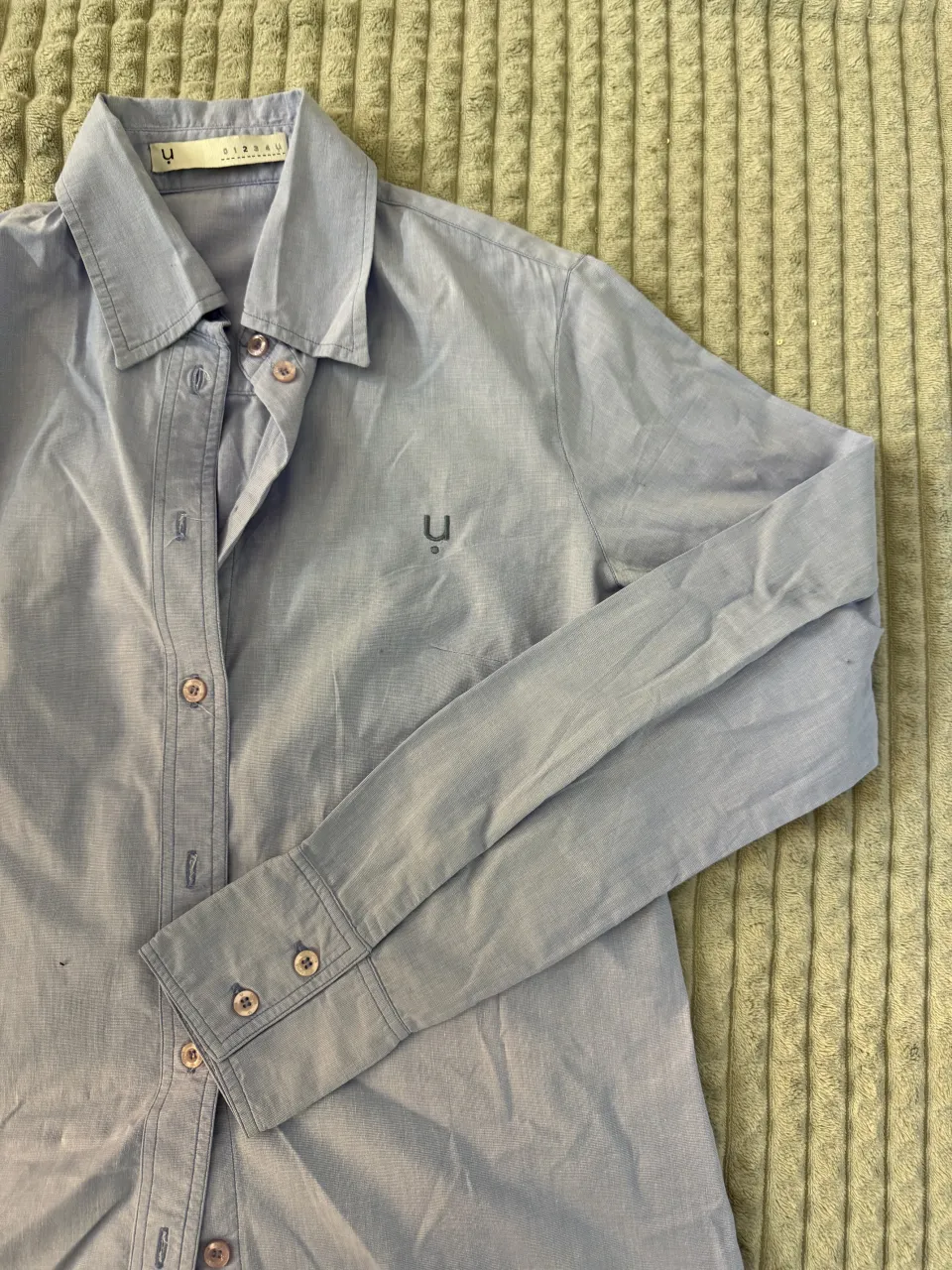 Camisa de manga larga color celeste con botones al frente y en los puños. Posee un pequeño logo bordado en el pecho.