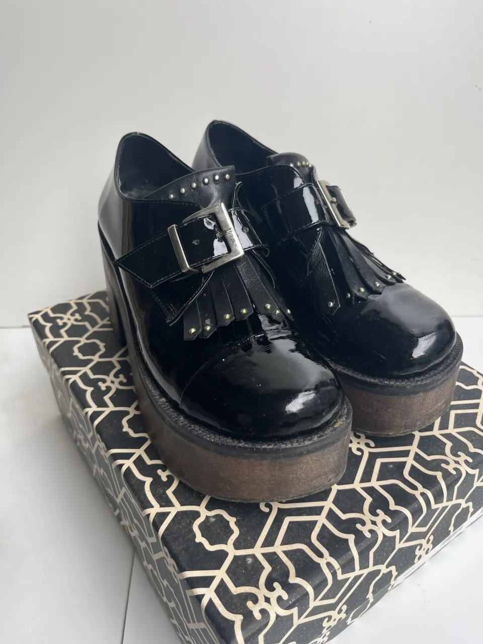 Zapatos negros de charol con plataforma y taco de madera. Tienen hebilla y flecos en la capellada. Detalle de tachas alrededor del cuello.