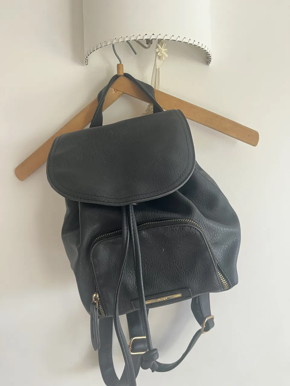 Mochila negra de cuero sintético con tapa y cordón ajustable. Posee un bolsillo frontal con cierre y tiradores dorados. Las correas son ajustables y también de cuero sintético.