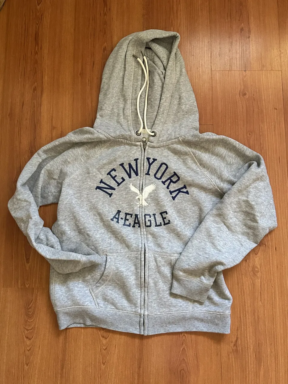 Buzo gris con capucha y cierre completo. Estampado en el frente con "NEW YORK" y un águila.